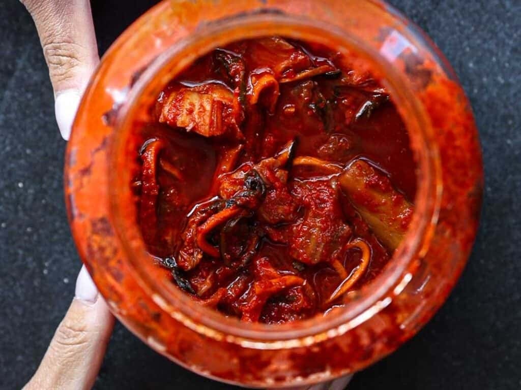 Open jar filled with red, spicy kimchi, showcasing pieces of fermented vegetables. Two hands hold the jar on a dark surface, highlighting the rich, vibrant colors and textures.